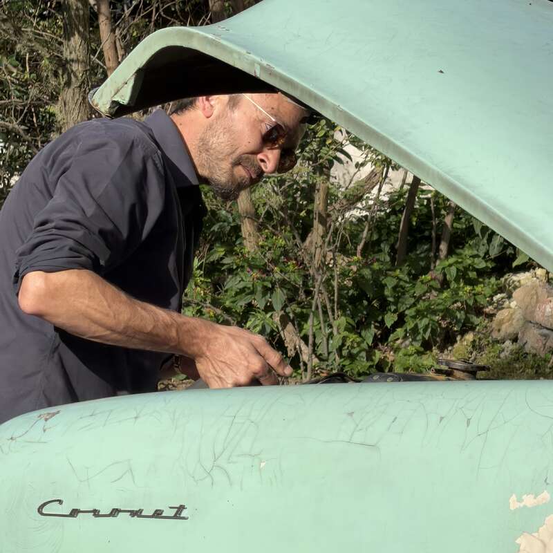 Ein Mann mit Sonnenbrille untersucht im Freien den Motorraum eines grünen Oldtimers, dessen Motorhaube geöffnet ist. An der Front ist der Modellname des Autos, „Coronet“, zu sehen.