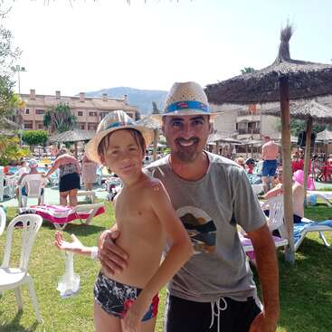 The image depicts a man and a boy posing for a photo at a resort, with the man wearing a hat and the boy in swimwear, set against a backdrop of a pool and other people.