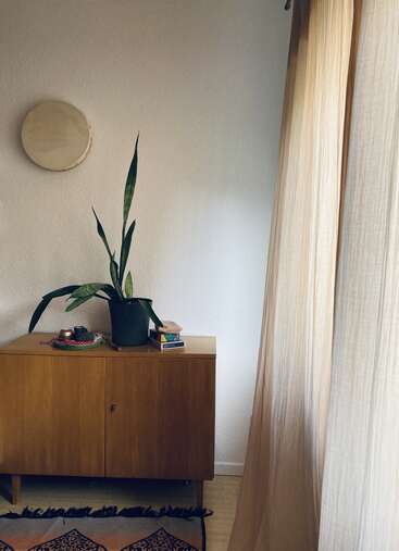 Eine minimalistische Zimmerecke mit einem Holzschrank, einer hohen Topfpflanze, Büchern, Kerzen, runder Wanddekoration, einem gemusterten Teppich und weichen beigen Vorhängen, die Licht hereinlassen.