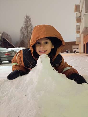 Un niño sonriente con una chaqueta naranja con capucha juega fuera en la nieve por la noche, arrodillado detrás de un pequeño montículo de nieve en una zona urbana nevada.