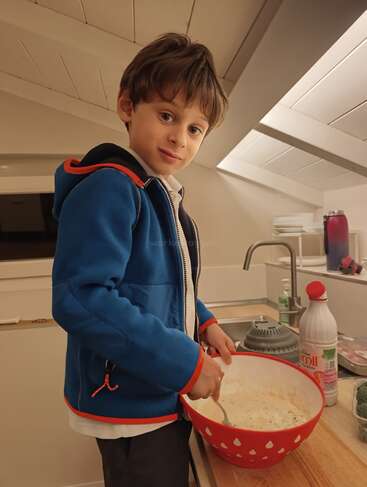 Un chico joven con chaqueta azul está de pie en una cocina, mezclando masa en un gran cuenco rojo, sonriendo ligeramente. Le rodean ingredientes y utensilios de cocina.
