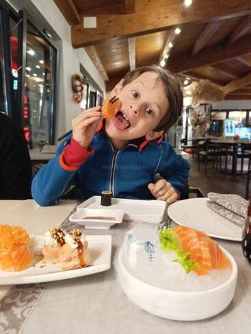 Un niño con chaqueta azul sostiene juguetonamente un trozo de sashimi de salmón con palillos, rodeado de platos variados de sushi en un acogedor restaurante.