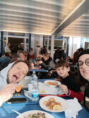 Una alegre familia disfruta de una mariscada en un concurrido restaurante informal. La gente cena a su alrededor y la luz del sol llena de calidez el animado ambiente.