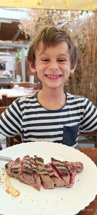 Un joven sonriente con camisa de rayas se sienta a la mesa de un restaurante y posa entusiasmado con un plato de filete a la parrilla delante.