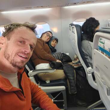 Un hombre se hace un selfie en un avión. A su lado, una mujer y un niño sonríen, mientras otro pasajero se sienta en la fila de delante, mirando hacia delante.