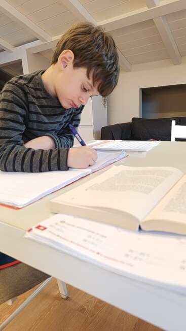 Un niño está sentado a la mesa, concentrado en escribir en un cuaderno, rodeado de libros y papeles abiertos, en un acogedor ambiente hogareño.