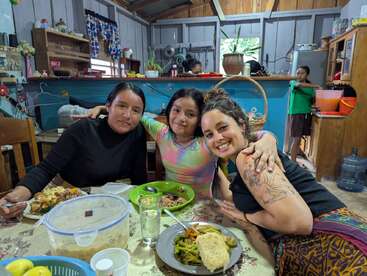 Three women sit closely together, smiling at the camera, enjoying a meal in a cozy, rustic kitchen. Two people are visible in the background, preparing food.