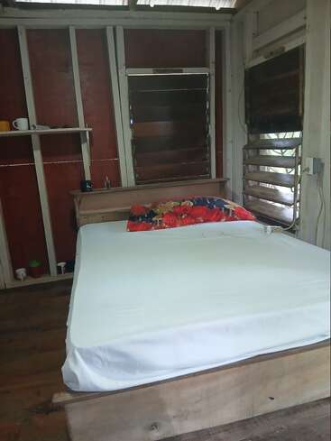 A simple rustic bedroom with a wooden bed, white sheet, colorful pillow, wooden floor, red and white walls, louvered windows, and a small shelf.
