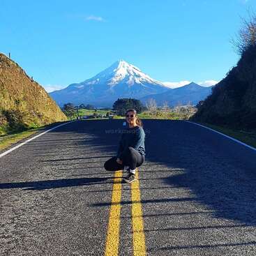 Une femme est assise sur une route, avec une majestueuse montagne enneigée en arrière-plan, sur fond de ciel bleu clair par une journée ensoleillée.