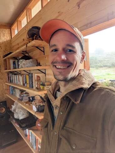 Ein lächelnder Mann in einer braunen Jacke und einer orangefarbenen Mütze steht in einem gemütlichen Holzraum mit Regalen voller Bücher und sieht neben einem Fenster fröhlich aus.