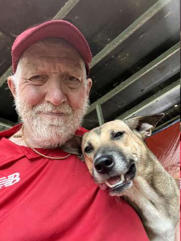 A smiling elderly man in a red shirt and cap takes a cheerful selfie with a happy, light-brown dog, both looking content and relaxed together.