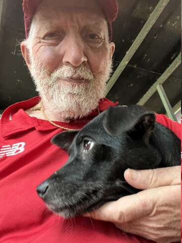 An elderly man with a white beard, wearing a red shirt and hat, smiles warmly while holding a small black dog close to his chest.