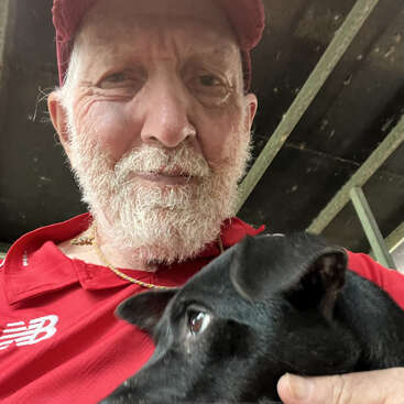 A bearded man wearing a red shirt and cap is holding a small black dog close to his chest, both appearing relaxed and comfortable together.