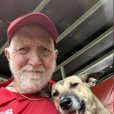Un sonriente anciano con camisa roja y gorra se toma un alegre selfie con un feliz perro marrón claro, ambos parecen contentos y relajados juntos.
