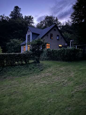 A cozy house with glowing windows sits surrounded by lush greenery and trees. The evening sky is dusky, creating a peaceful and inviting atmosphere.