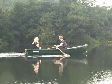 A man rows a green boat across a calm lake, accompanied by a blonde woman. Lush green trees reflect on the peaceful, mirror-like water surface.