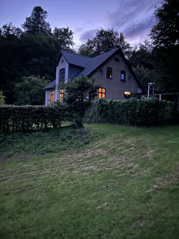 A cozy house with glowing windows sits on a lush, green lawn surrounded by trees at dusk, creating a peaceful and inviting atmosphere.