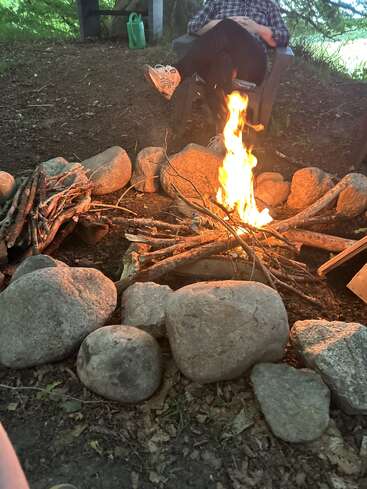 A cozy campfire burns within a circle of large rocks, surrounded by sticks, with a person relaxing in a chair nearby amidst a forest setting.