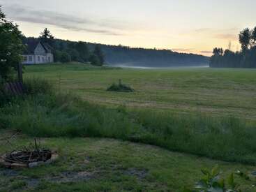 The image depicts a serene landscape featuring a house, lush green field, and trees, with a small fire pit in the foreground, set against a vibrant sunset sky.