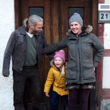Das Bild zeigt eine dreiköpfige Familie, die vor einer Holztür steht, mit dem Vater auf der linken Seite, der Tochter in der Mitte und der Mutter auf der rechten Seite.