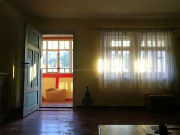 The image depicts a room with an open door leading to a porch, featuring a window with white curtains, and a wooden floor, bathed in sunlight.
