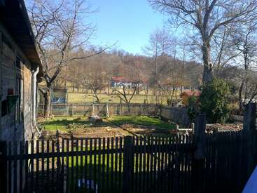 The image depicts a serene rural landscape featuring a house, a wooden fence, and a garden, with trees and a clear blue sky in the background.