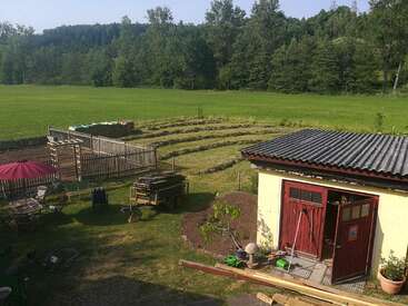 The image depicts a serene rural scene, featuring a small yellow shed with a red door, surrounded by lush greenery and a vibrant garden, set against a picturesque backdrop.