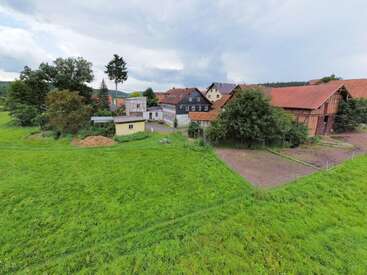 The image depicts a serene rural scene featuring a cluster of houses and barns surrounded by lush green fields and trees under a cloudy sky.