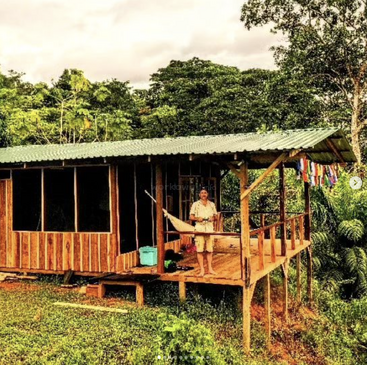 Un homme se tient sous le porche d'une cabane en bois au toit vert, entourée d'arbres et de feuillages luxuriants, dans un cadre extérieur serein.