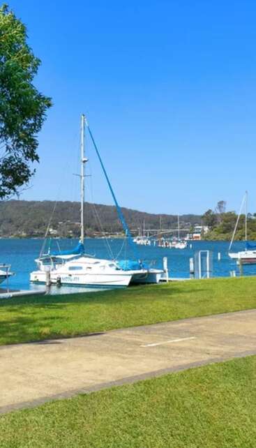 Eine friedliche Seeszene mit angedockten Segelbooten auf klarem, blauem Wasser, grünem Gras, einem Baum, entfernten Hügeln und einem hellen, wolkenlosen blauen Himmel.