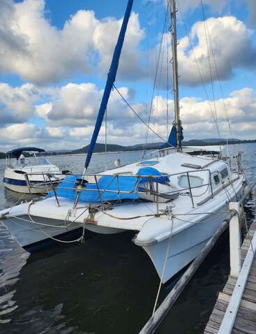 Ein weißes Katamaran-Segelboot liegt in einem Yachthafen, umgeben von ruhigem Wasser. Flauschige Wolken füllen den blauen Himmel, und in der Ferne sind die Hügel zu sehen. Eine friedliche Szene.
