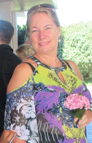 Das Bild zeigt eine Frau mit blonden Haaren, die ein gelbes Oberteil mit einem lila Muster trägt und an einem sonnigen Tag draußen steht, umgeben von üppigem Grün und anderen Menschen.