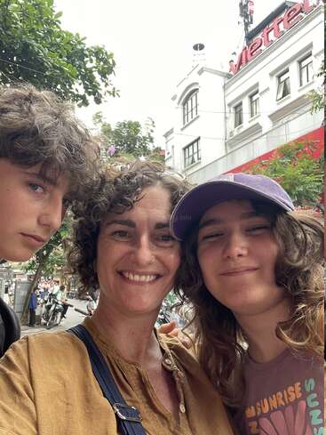 Três pessoas de cabelo cacheado sorriem para uma selfie na rua, cercadas por vegetação e prédios. Um grande letreiro com a palavra “Viettel” aparece ao fundo.