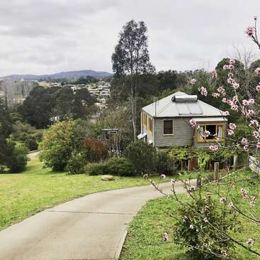 Uma casinha com detalhes em amarelo fica em meio a uma vegetação exuberante, árvores floridas e colinas onduladas, com vista para uma cidade tranquila sob um céu nublado e sereno.