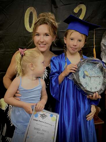 Una madre orgullosa posa con sus dos hijas en una ceremonia de graduación. Una de las hijas lleva toga y birrete azules y sostiene un reloj y un diploma.