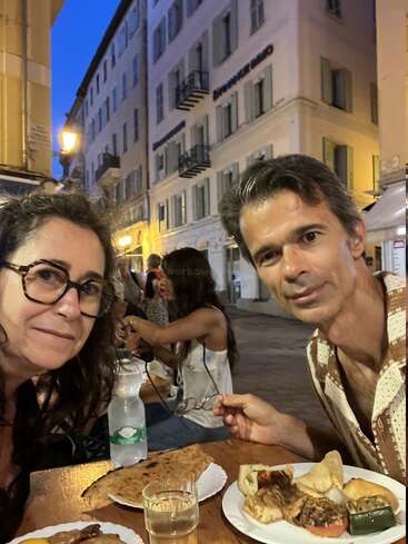 Un hombre y una mujer cenan al aire libre por la noche en una encantadora calle europea, disfrutando de platos de comida y bebidas con edificios históricos de fondo. Ambiente acogedor.
