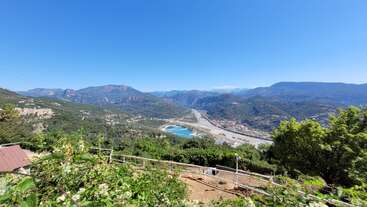 Esta imagen muestra un pintoresco paisaje de montaña bajo un cielo azul despejado, con exuberante vegetación, una pequeña zona vallada, un río y un embalse azul en el valle.