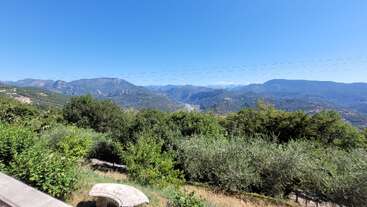 Esta imagen muestra un pintoresco paisaje de montaña bajo un cielo azul despejado, con frondosos árboles y arbustos verdes en primer plano y un banco de piedra.