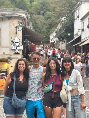 Cuatro personas posan felices en una bulliciosa calle del casco antiguo. El fondo tiene mucha gente, tiendas, restaurantes y vegetación. Todos parecen relajados, disfrutando juntos de un día de verano.