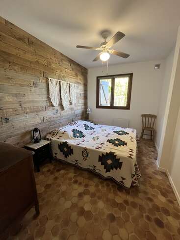 Este acogedor dormitorio cuenta con una pared rústica de madera, una cama de matrimonio con una funda de motivos geométricos, ventilador de techo, silla de madera, ventana y suelo de tonos cálidos.