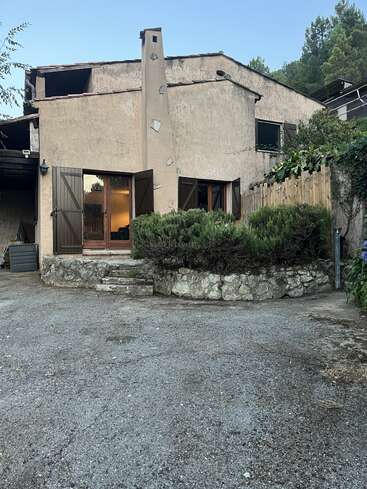 Una casa rústica con una chimenea única, escalones de piedra, contraventanas de madera y un camino de entrada de grava. Rodeada de vegetación y árboles, parece tranquila y acogedora.
