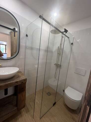 Este moderno cuarto de baño cuenta con una ducha acristalada, un inodoro montado en la pared, un tocador de madera rústica con lavabo de recipiente, un espejo redondo y suelo de madera clara. Diseño luminoso, minimalista y elegante.