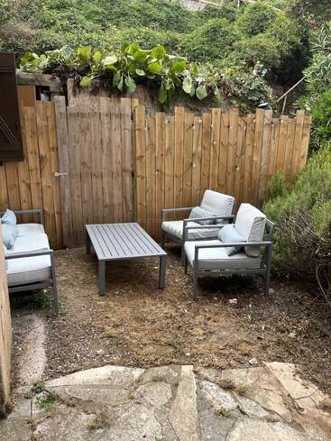 Un pequeño patio con muebles de exterior grises: una mesa de centro, dos sillas y un sofá de dos plazas. Una valla de madera, luces de cuerda y una frondosa vegetación rodean la acogedora zona.