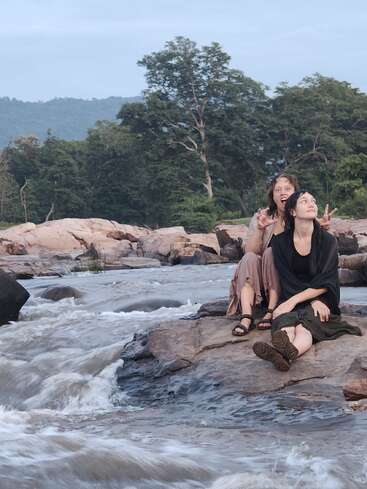 Two women sit on rocks by a flowing river, surrounded by lush greenery and trees. One makes a playful gesture; both appear relaxed and happy.