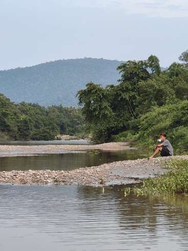 A young man sits thoughtfully on a rocky riverbank, wearing a mask, surrounded by lush green trees, calm water, and distant forested hills. Peaceful scene.