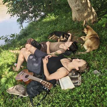 Two people relax on grass under a tree beside a river, accompanied by two dogs. A guitar, book, and hat lie nearby, suggesting peaceful leisure.