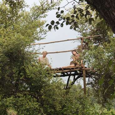Two people sit on a simple wooden platform built high among tree branches, surrounded by lush green foliage, enjoying peaceful nature and a scenic, elevated view.
