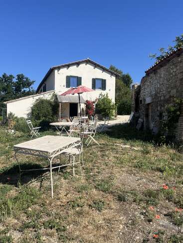 Une charmante maison de campagne aux murs blancs et aux volets verts se détache sous un ciel bleu limpide. À l'extérieur, on trouve un coin salon avec des tables et des chaises en fer forgé, ainsi qu'un parasol rouge.