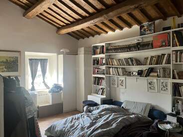 Une chambre chaleureuse avec un plafond rustique aux poutres apparentes, un lit défait, des étagères blanches remplies de livres, une fenêtre ornée de rideaux bleus et des touches décoratives personnelles.