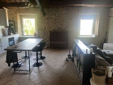 Cette pièce chaleureuse et rustique est dotée de murs en pierre, de deux grandes fenêtres, d'une cuisine moderne, d'une table à manger avec des chaises, d'un canapé, d'une bibliothèque et d'une lumière naturelle qui inonde l'espace.